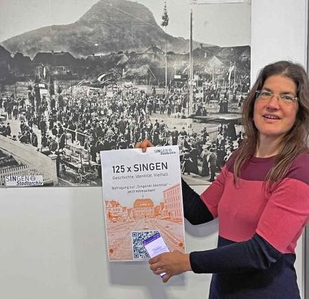 Stadtarchivarin Britta Panzer mit dem Plakat zur Online-Befragung. Im Hintergrund ist ein Foto der Feierlichkeiten anlässlich der Stadterhebung1899 zu sehen. 